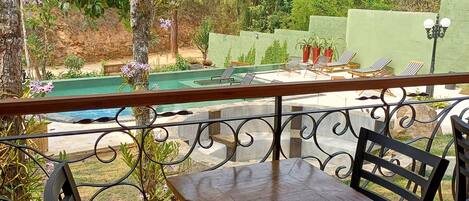 Un área de estar exterior techada con vista a la piscina, que incluye una mesa y sillas de madera, rodeada de follaje y una pared verde.