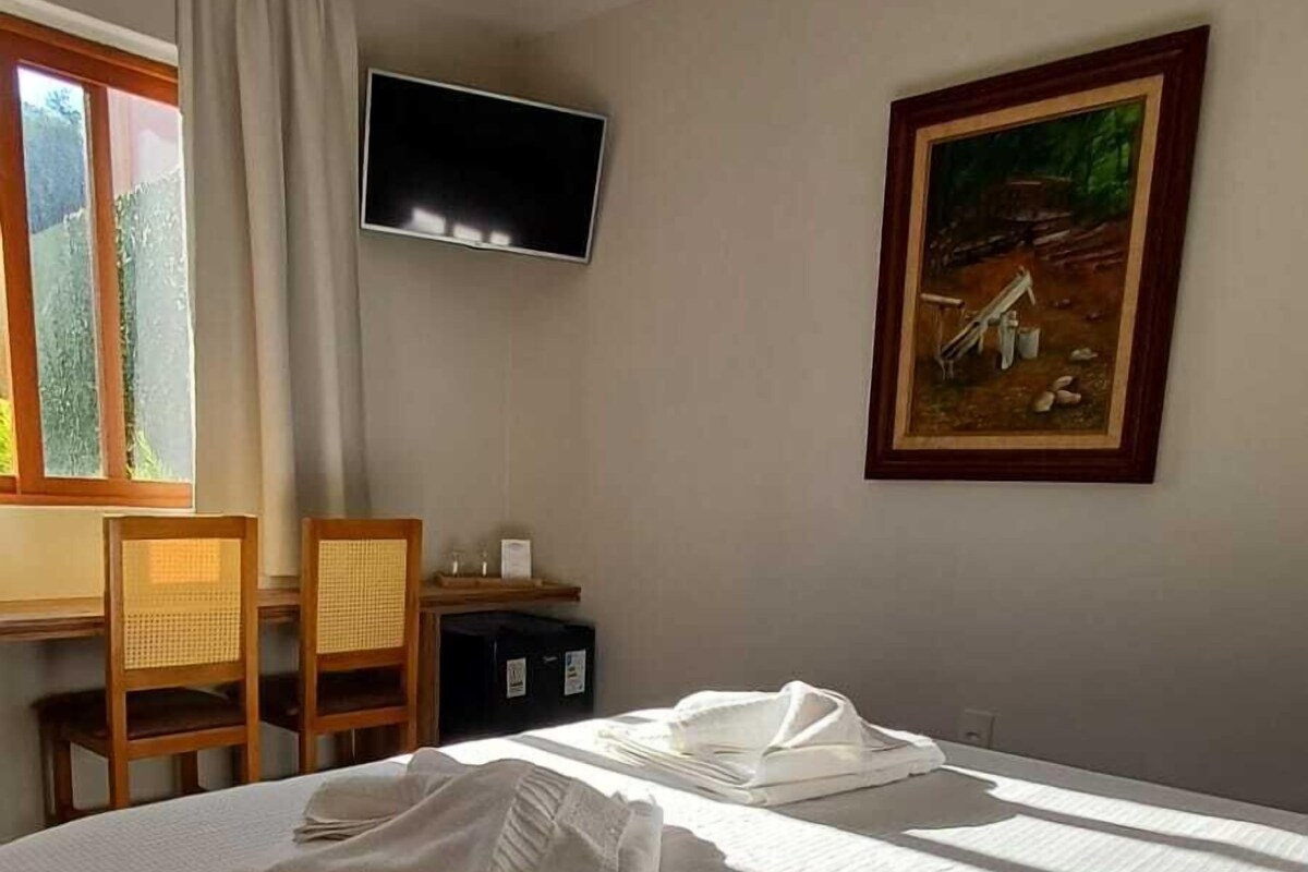 A hotel room with a bed, a desk, a chair, a television, and a painting on the wall.