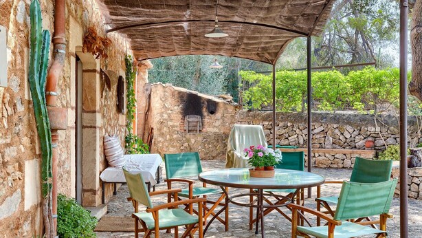 Une terrasse extérieure avec une table ronde et des chaises, un espace abrité doté d’un mur en pierre rustique, et un cactus.