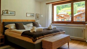 Un dormitorio con una cama grande, una ventana que ofrece vistas a una montaña y edificios, un radiador y una mesita de noche con una lámpara.