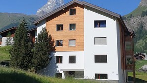 Un edificio de dos plantas con fachada de madera y numerosas ventanas, sobre un fondo de montañas y cielo despejado.