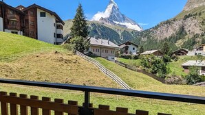 Un pueblo de montaña con casas en una ladera, un pico montañoso destacado al fondo y una valla de madera en primer plano.