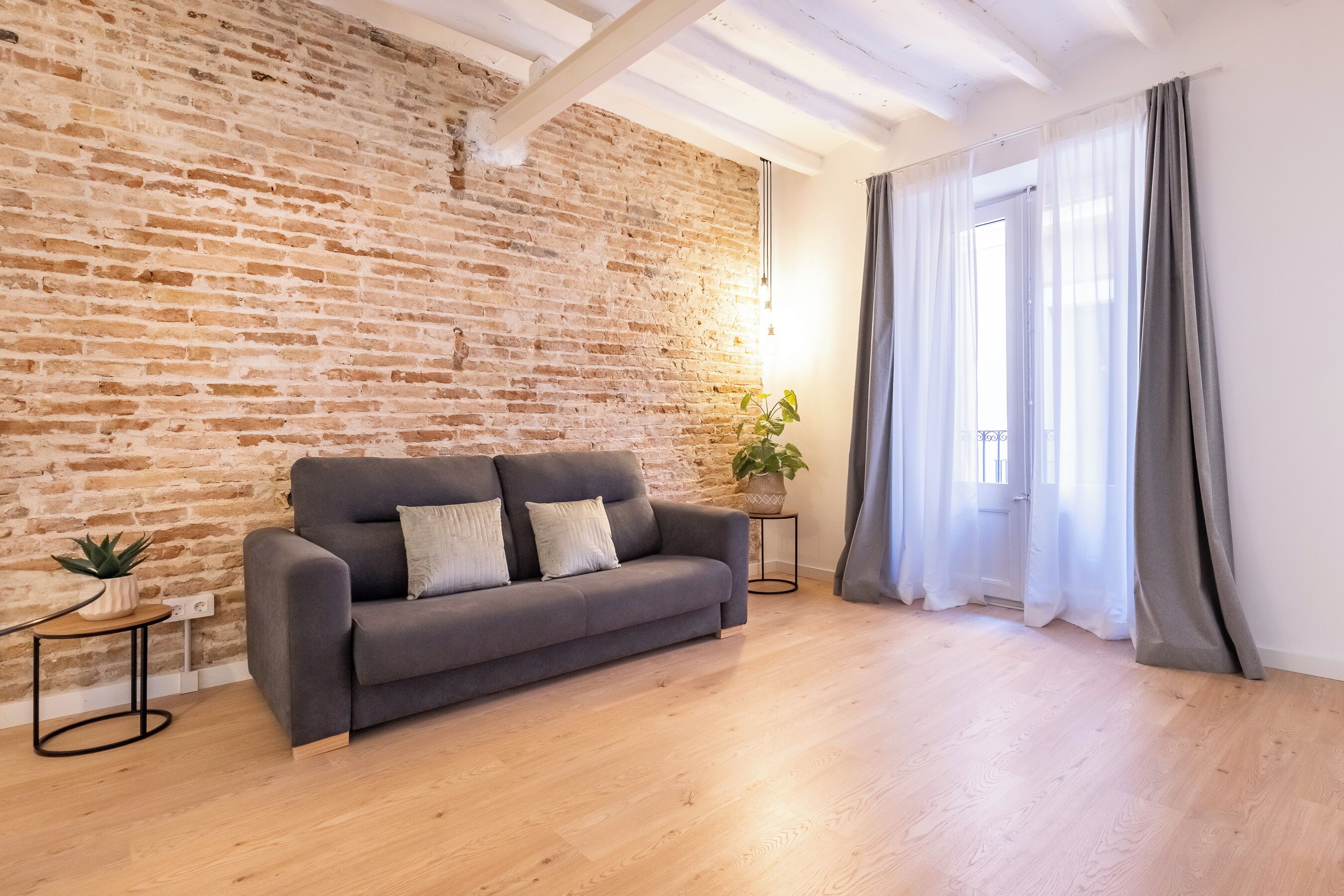 Una sala de estar con pared de ladrillo, un sofá gris, piso de madera, una mesita con una planta y una ventana con cortinas.
