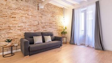 Una sala de estar con pared de ladrillo, un sofá gris, piso de madera, una mesita con una planta y una ventana con cortinas.