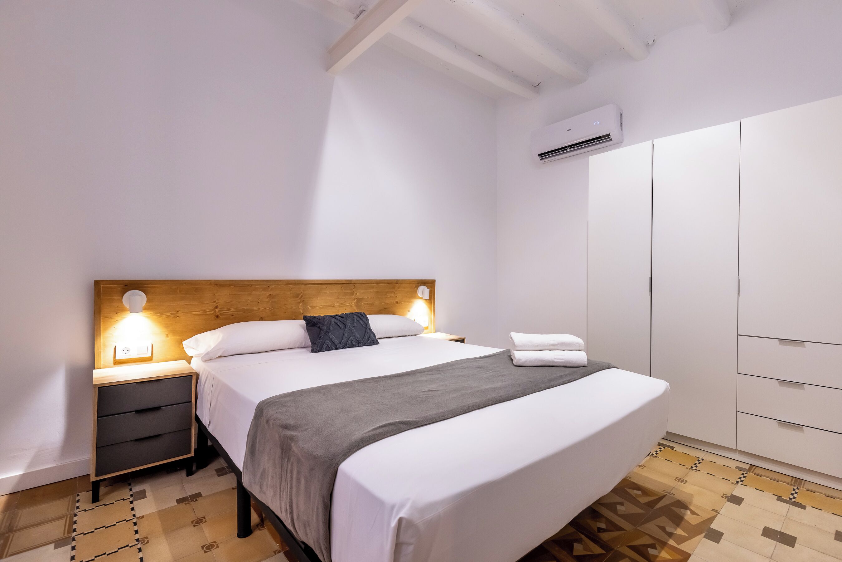 A neatly made bed with a wooden headboard, a bedside table with a lamp, and a wardrobe in a well-lit room.