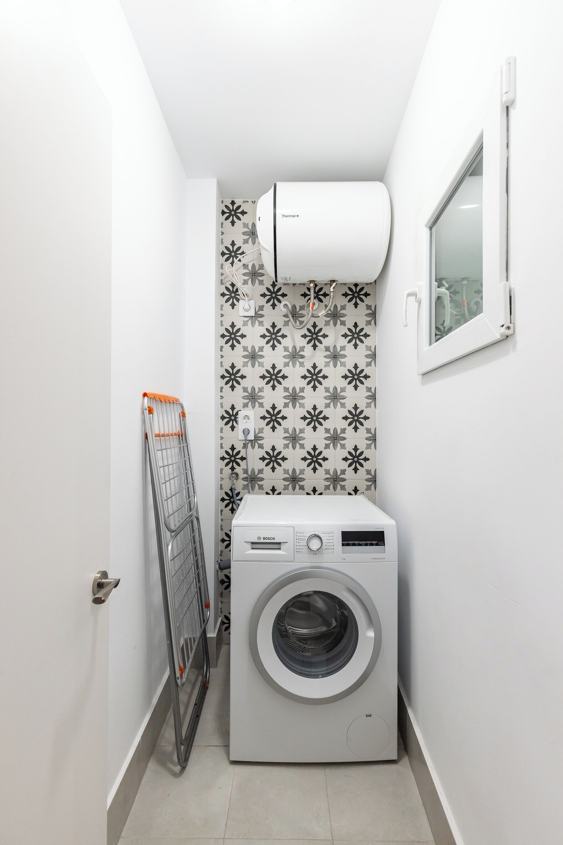 A compact laundry room with a Bosch washing machine, a drying rack, and a Bosch Thermostatic Air Conditioner mounted on the wall.