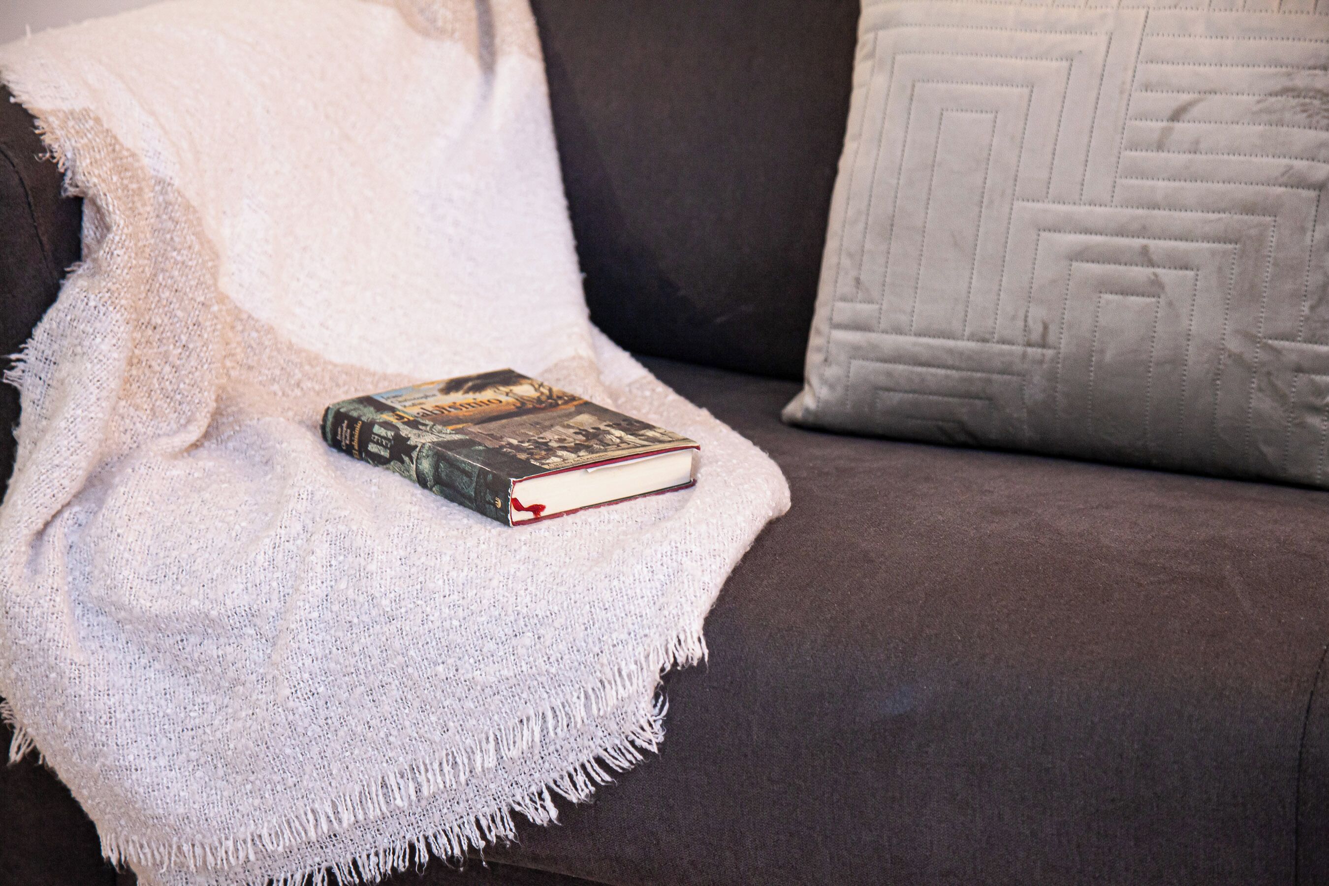 Ein Buch auf einer Couch mit einer weißen Decke und einem gemusterten Kissen.