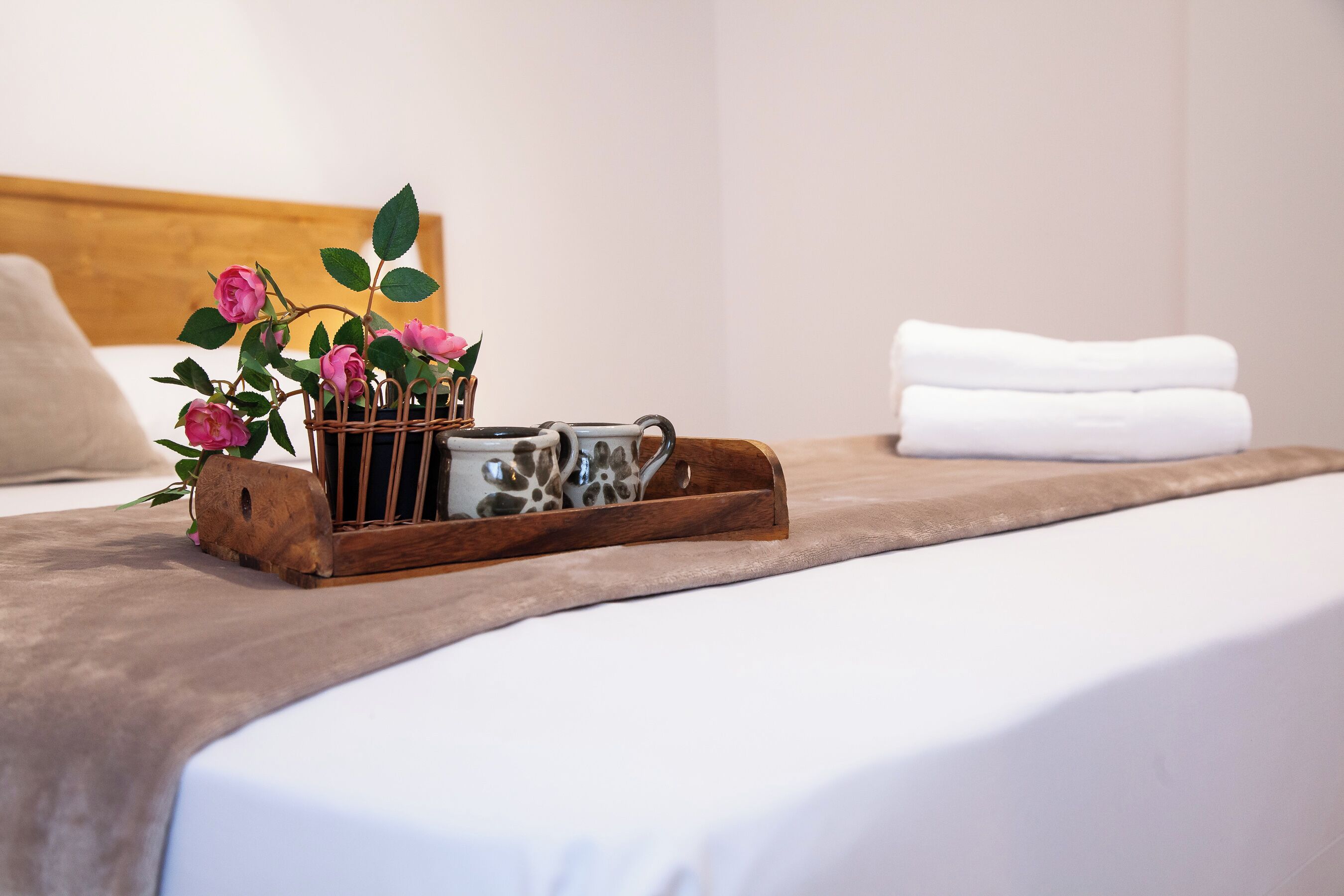 A neatly made bed with a brown bedspread, a wooden tray holding a camera and pink flowers, and a white pillow in a hotel room.