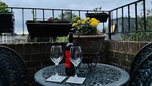 Une table ronde en métal, agrémentée de deux verres à vin et d’une bouteille, est entourée de chaises noires décorées. L’ensemble est installé sur un balcon orné de plantes en pot et muni d’une balustrade.