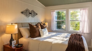 A bedroom with a bed, nightstand, lamp, decorative wall piece, and windows with curtains. - 1880s Schoolhouse/Hot Tub/Firepit/Hocking Hills (McArthur)