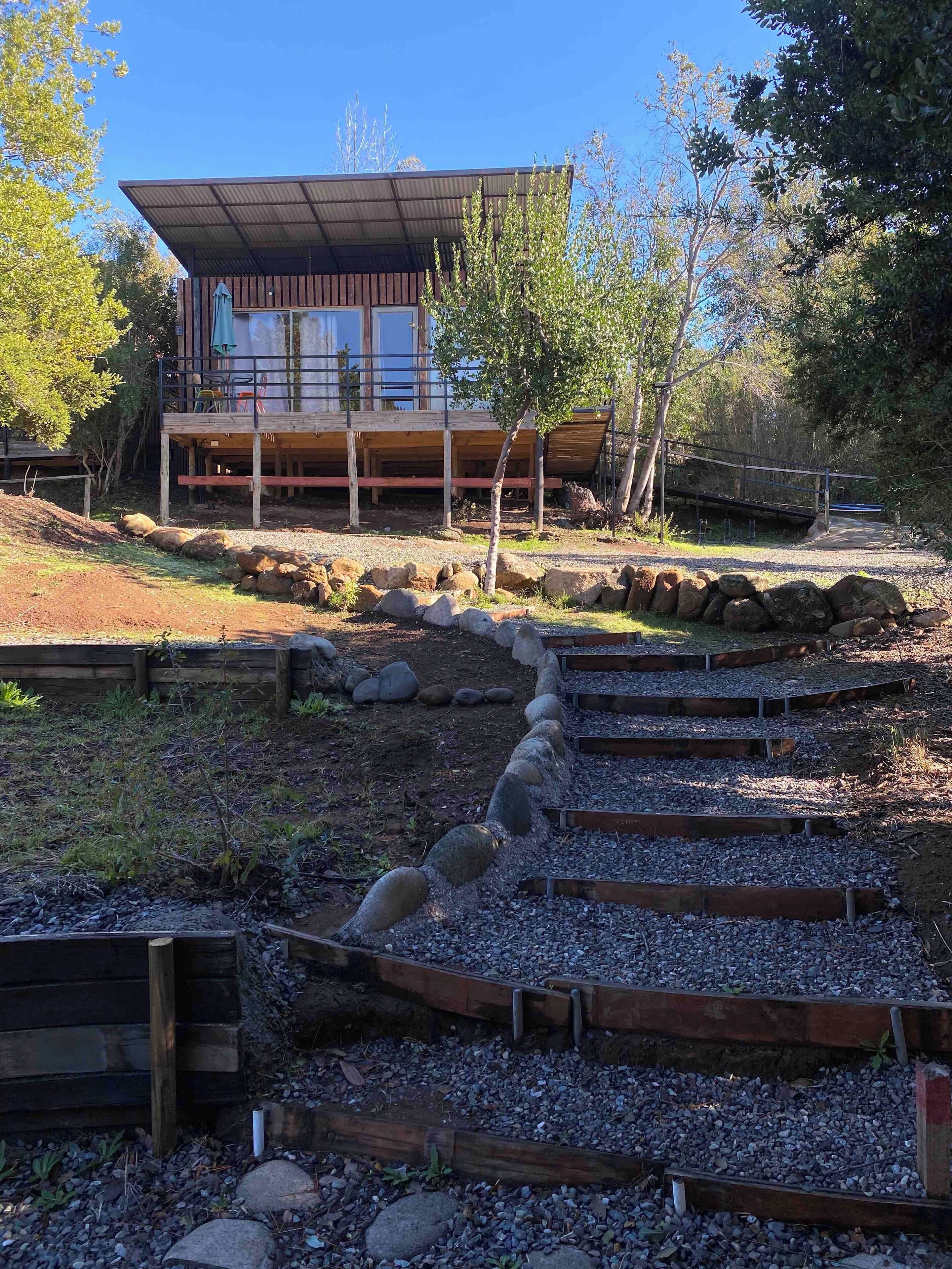 A modern building with a flat roof and large windows, surrounded by a gravel pathway and wooden steps leading to a wooden deck with chairs.