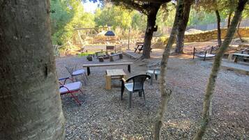 An outdoor seating area with tables and chairs, surrounded by trees and a stone wall.