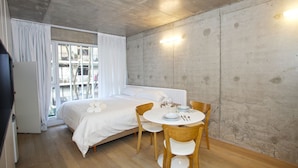 Een slaapkamer met een bed, een eettafel met stoelen, een raam met gordijnen en een betonnen muur.