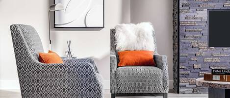 Dos sillones tapizados con estampado y cojines naranjas, un cuadro abstracto moderno, una pared decorativa con diseño de ladrillo y una mesita con un letrero.