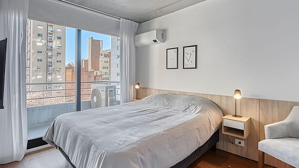 Ein modernes Schlafzimmer mit einem großen Bett, einem hölzernen Kopfteil, einem Nachttisch, einem Sessel und Blick auf Gebäude durch ein großes Fenster.