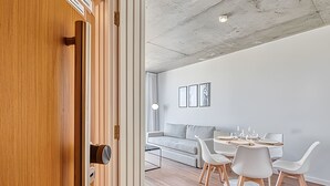 Een modern interieur met een houten deur, een grijze bank, een ronde tafel met stoelen en kunstwerken in lijsten aan de muur.