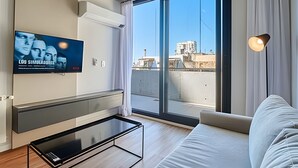 Een moderne woonkamer met een flatscreen televisie aan de muur, een glazen salontafel, een bank en een groot raam met uitzicht op de stad.