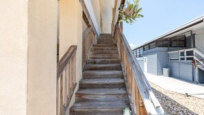 Un escalier en bois mène à un bâtiment à la façade blanche, orné d’une plante suspendue.