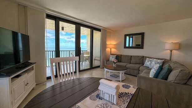 A living room with a TV, a sofa, a coffee table, and a view of the sea through large glass doors. - Sea Winds 1401: Beach Front; Pool View (Marco Island)