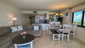 A modern living room with a sofa, coffee table, and dining area with chairs. A kitchen is visible in the background. - Sea Winds 1401: Beach Front; Pool View (Marco Island)