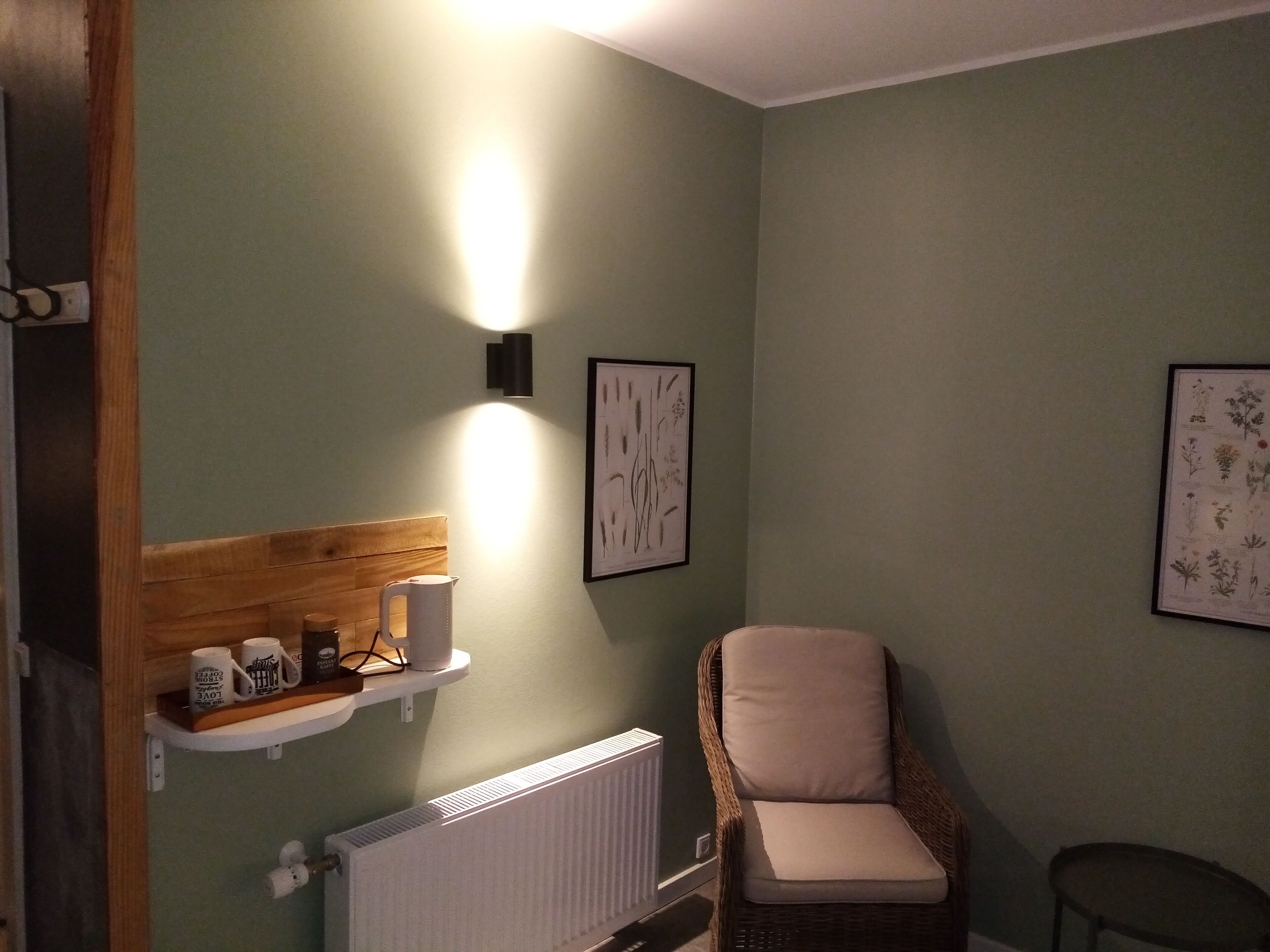 A room with a wicker chair, a radiator, a shelf with mugs, and framed botanical prints on the wall.