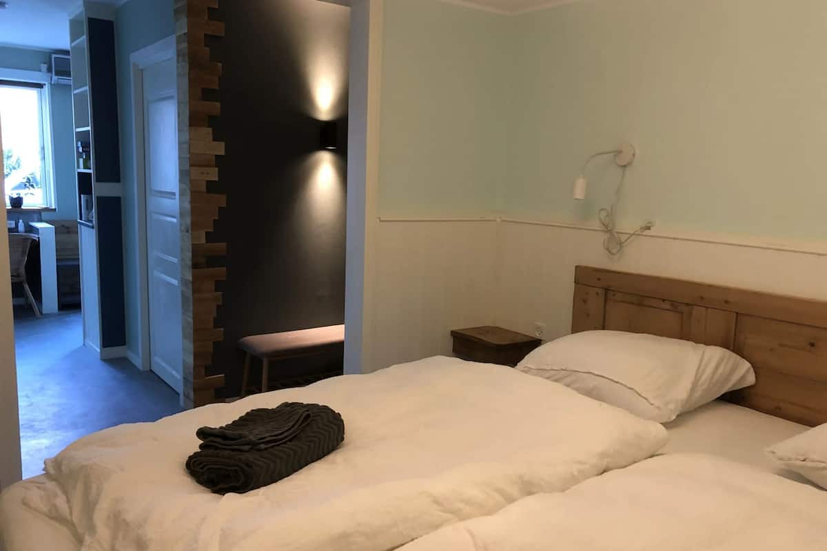 Ein Schlafzimmer mit einem Bett, einem Nachttisch, einem Sessel und einem kleinen Tisch. Eine Tür führt in einen anderen Raum.