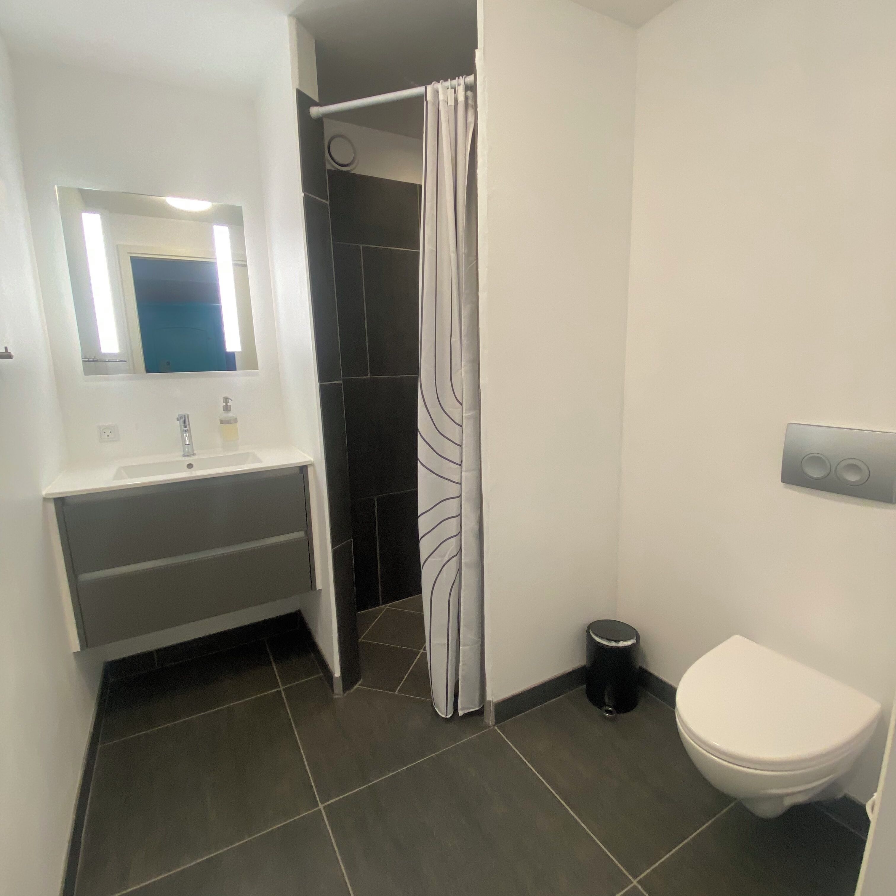 Un baño con lavamanos blanco, un espejo, cortina de ducha, inodoro y un cubo de basura.