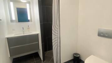 Un baño con lavamanos blanco, un espejo, cortina de ducha, inodoro y un cubo de basura.