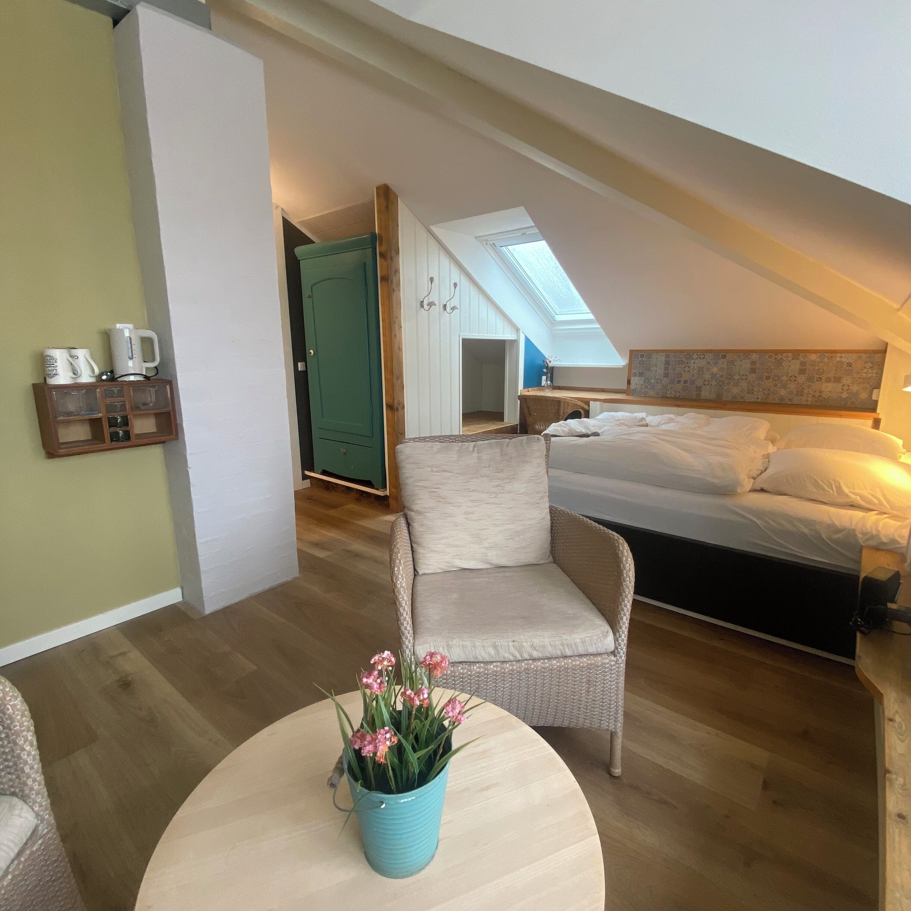 A cozy bedroom with a bed, a chair, a small table with flowers, a green door, and a skylight.