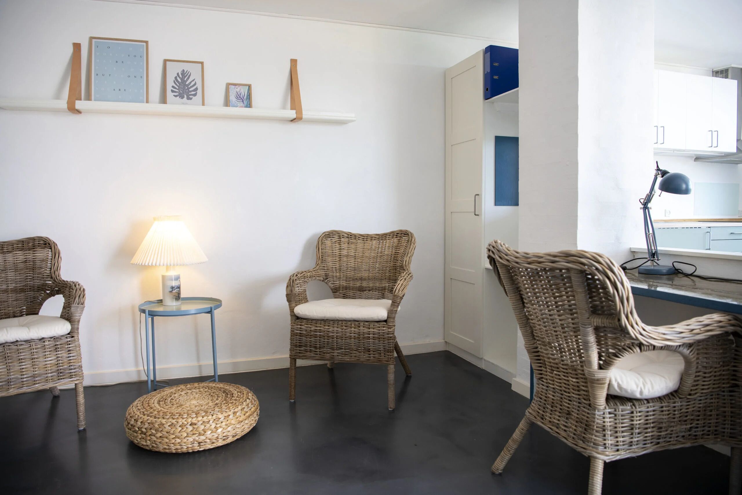 Una habitación con tres sillas de mimbre, un pequeño taburete redondo de mimbre, una mesita auxiliar azul con una lámpara y una estantería con cuadros enmarcados.