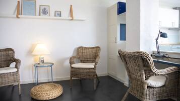 Una habitación con tres sillas de mimbre, un pequeño taburete redondo de mimbre, una mesita auxiliar azul con una lámpara y una estantería con cuadros enmarcados.
