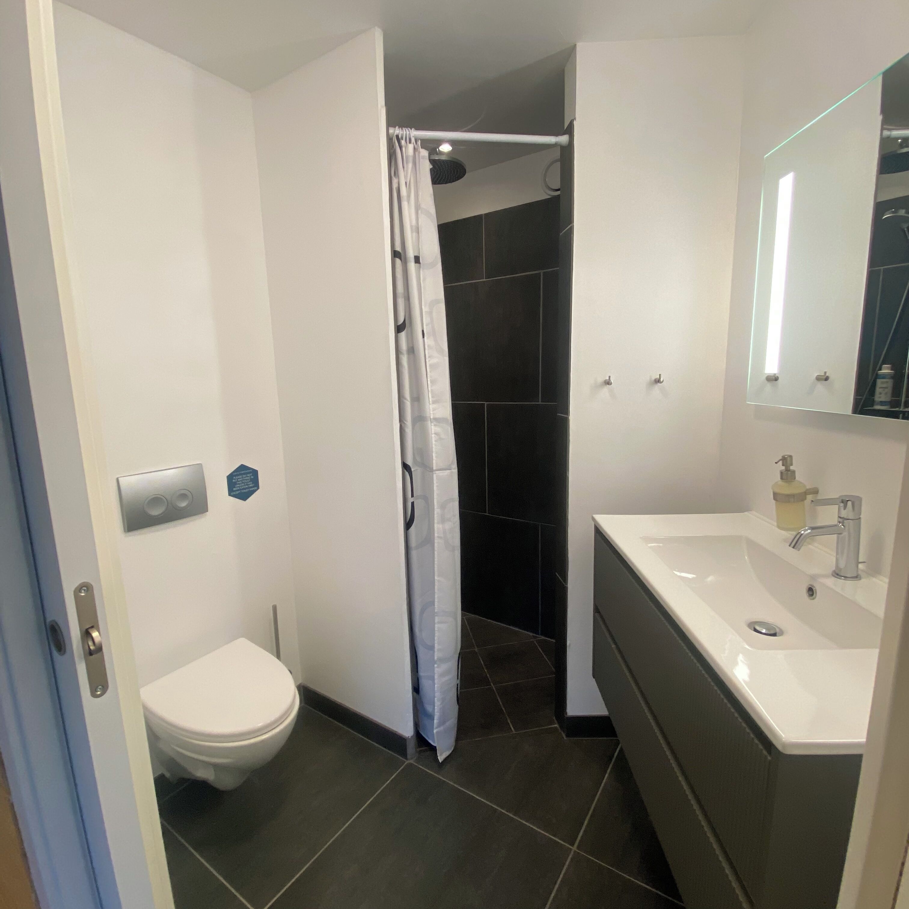 Een moderne badkamer met toilet, douche met gordijn, wastafel met spiegel en zeepdispenser.