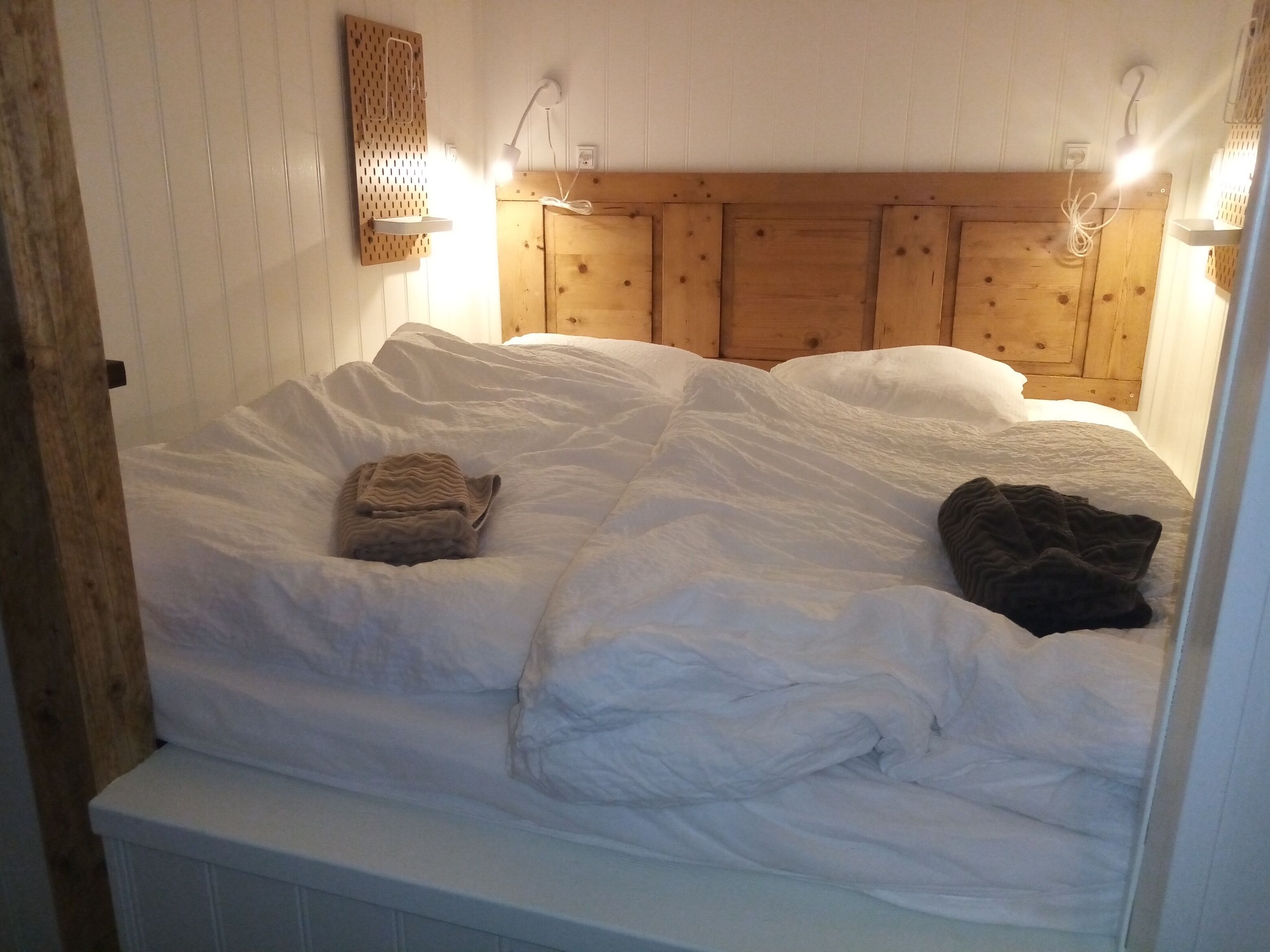 Una cama con ropa de cama blanca, dos almohadas y una colcha. Hay luces fijadas en la pared y un cabecero de madera.