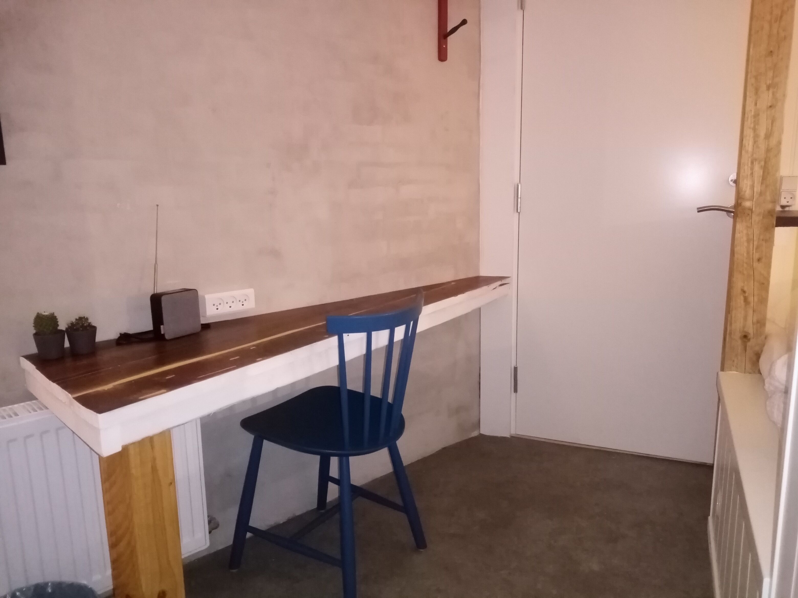 Una habitación pequeña y funcional con un escritorio de madera y una silla azul. Hay un radiador blanco debajo del escritorio y una puerta blanca al fondo.
