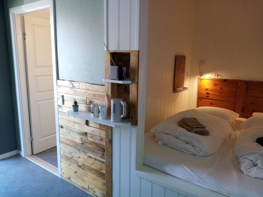 Quarto com cabeceira de madeira, cama com roupa de cama branca, mesa de cabeceira com chaleira e pequena prateleira com cactos e canecas.