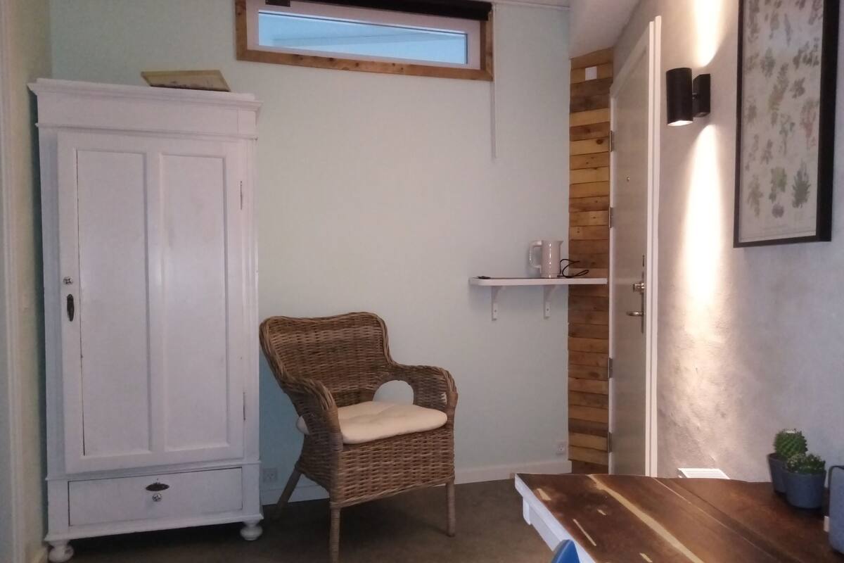 Ein Zimmer mit einem weißen Kleiderschrank, einem Korbstuhl, einem Holztisch, einer kleinen Ablage mit einer weißen Tasse und einem Fenster mit Holzrahmen.