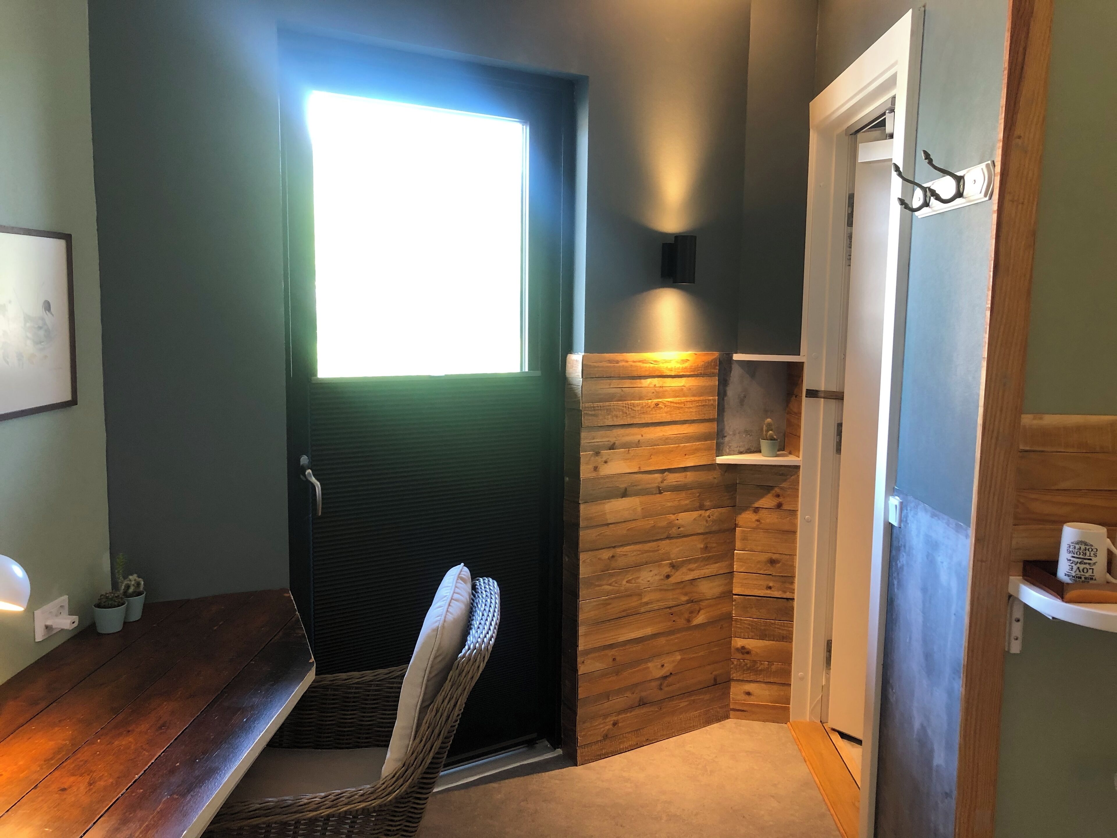 A room with a dark door, a small window, a wicker chair, a shelf with a cactus, and a wall-mounted light.