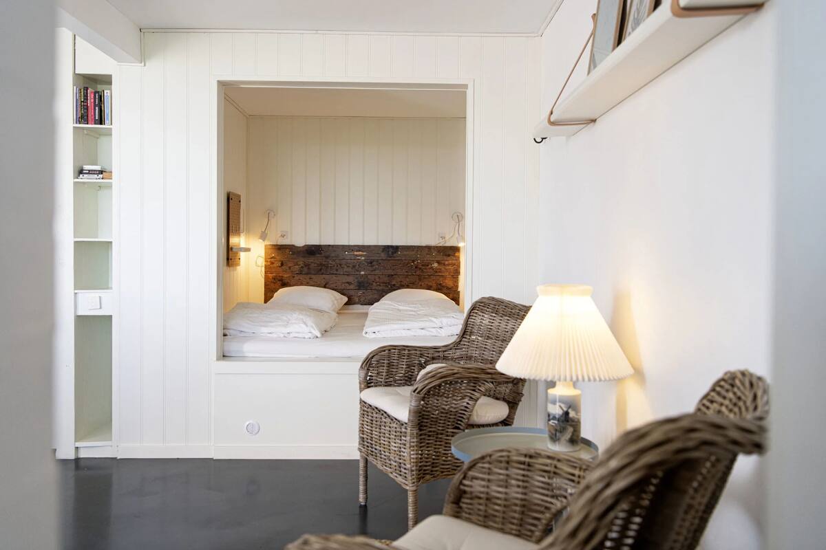 Ein gemütlicher Raum mit integriertem Bett, Korbmöbeln, einem kleinen Tisch mit Lampe und einem Bücherregal.