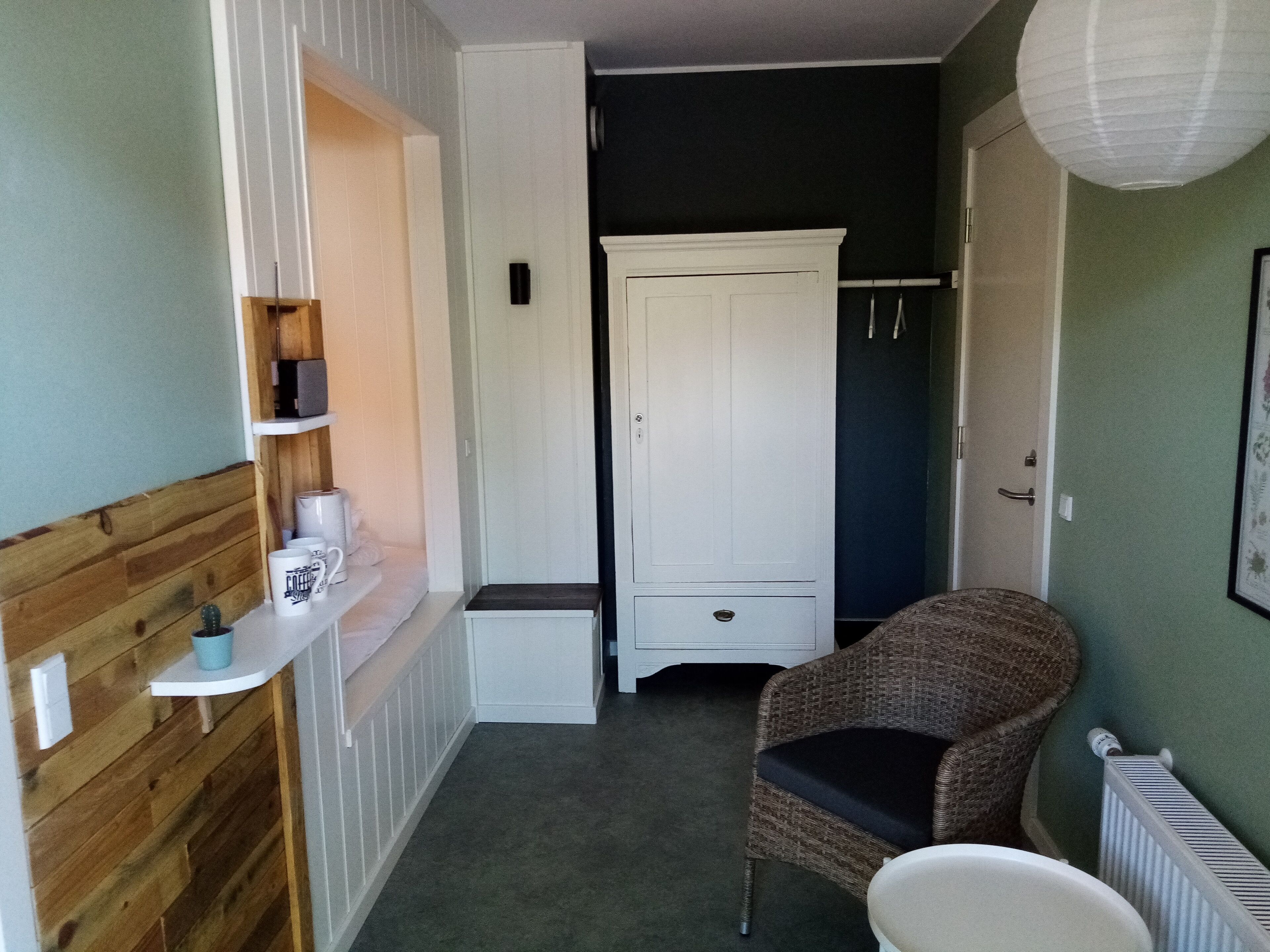 A small, narrow room with a wooden wall, a white wardrobe, a wicker chair, and a radiator.