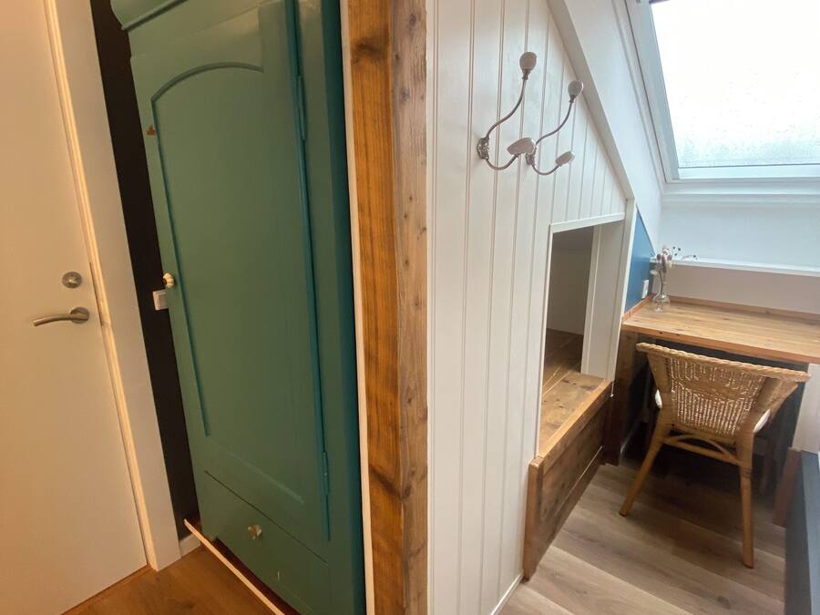 A room with a teal door, a wooden desk, a wicker chair, and a window with a view.