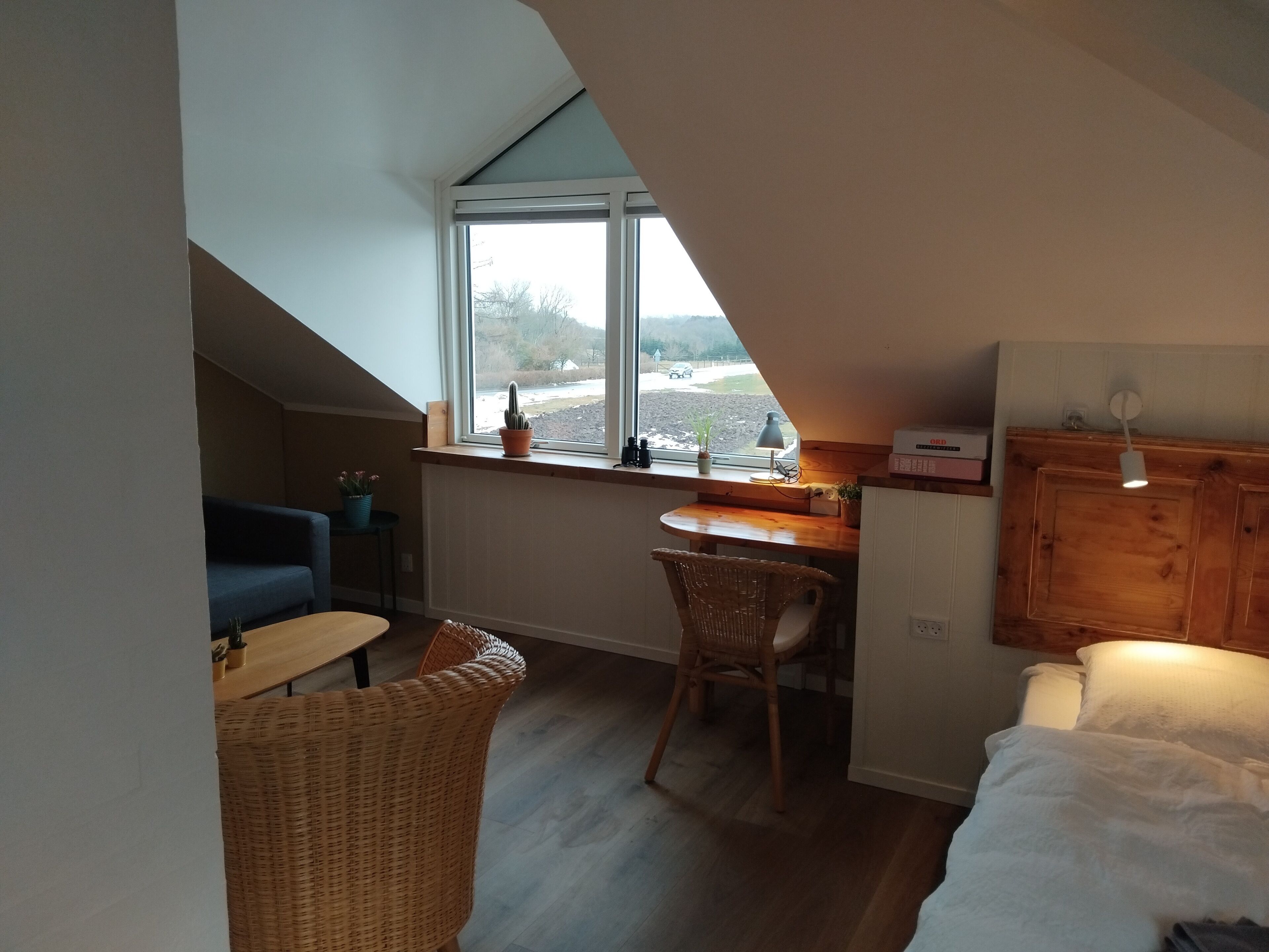 A cozy room with a sloped ceiling, a wooden desk, a wicker chair, a bed, and a window with a view of the outdoors.