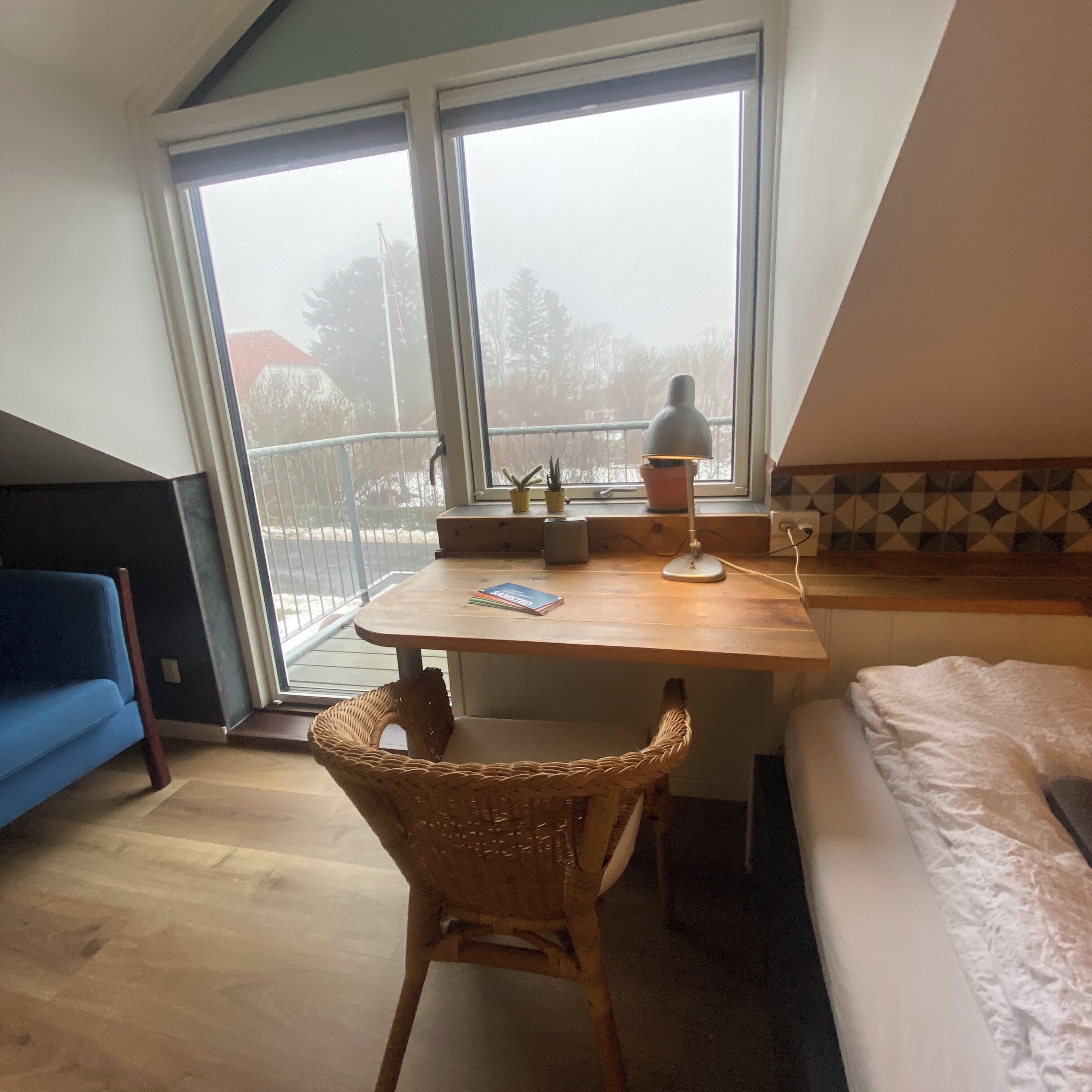 Una habitación con un escritorio de madera, una silla de mimbre, una cama y un ventanal con vistas a árboles y un edificio.