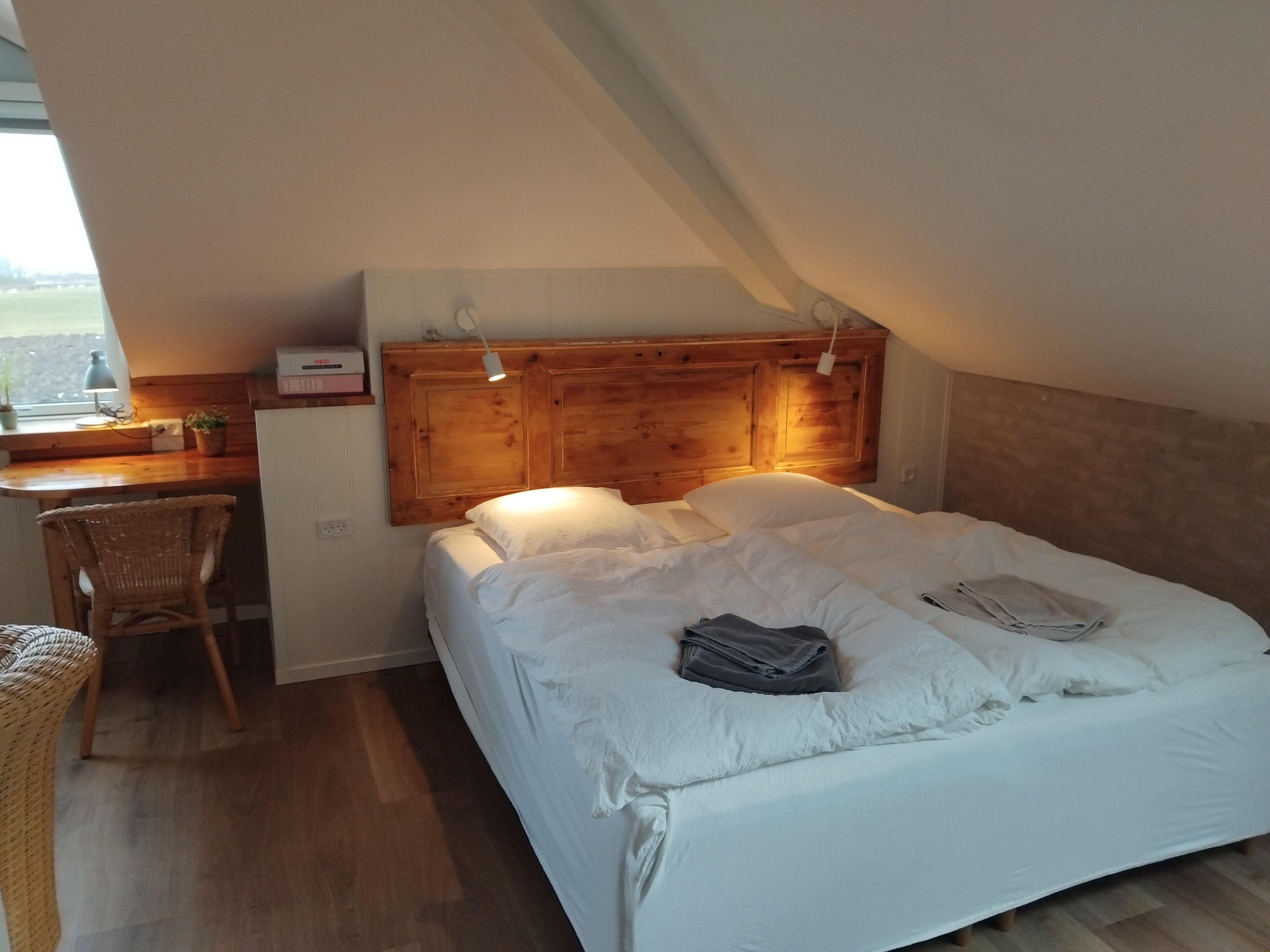 Una camera da letto con una testiera in legno, una scrivania con una lampada, una sedia in vimini e un letto con biancheria da letto bianca.