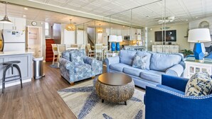 A modern living room with a blue sofa, patterned armchairs, a round coffee table, and a kitchen in the background. - Ocean Mile I-4 - Beach Front, Swimming Pool, Beach Gear Included! (St George Island)