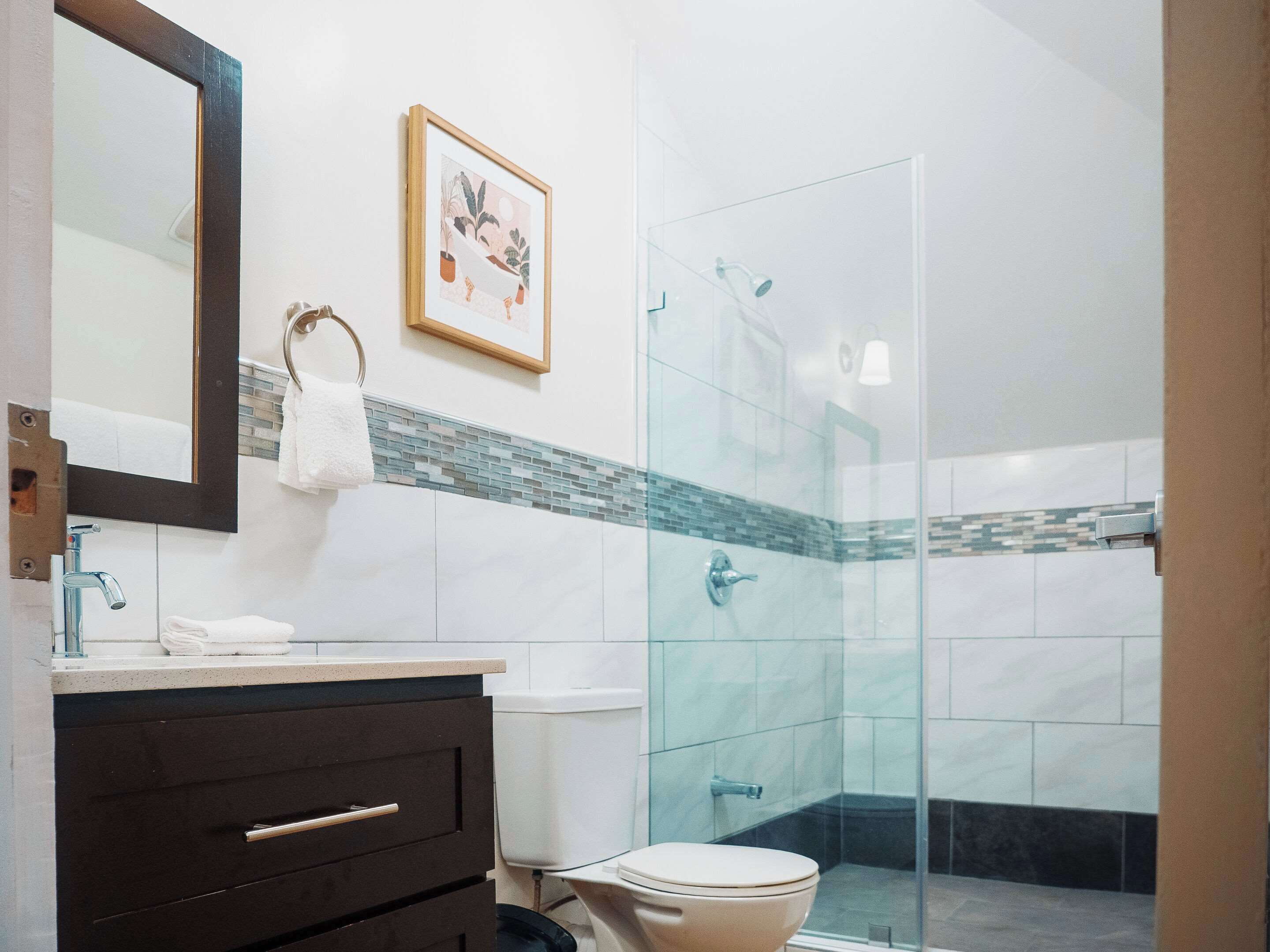 A modern bathroom with a glass shower enclosure, a white toilet, a vanity with a mirror, and a framed picture on the wall.