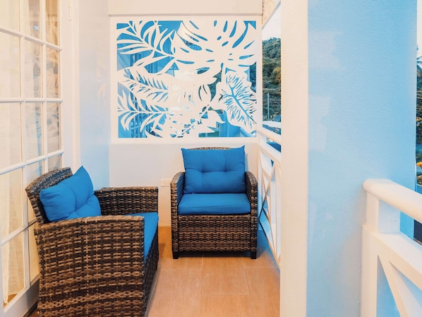 Ein Balkon mit Korbmöbeln und blauen Kissen, ein dekoratives Blattmuster an der Wand und Blick ins Freie.