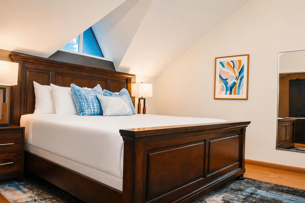 Une chambre à coucher avec un grand lit, une tête de lit en bois, des lampes de chevet, un tableau abstrait aux couleurs bleu et blanc, et une lucarne.
