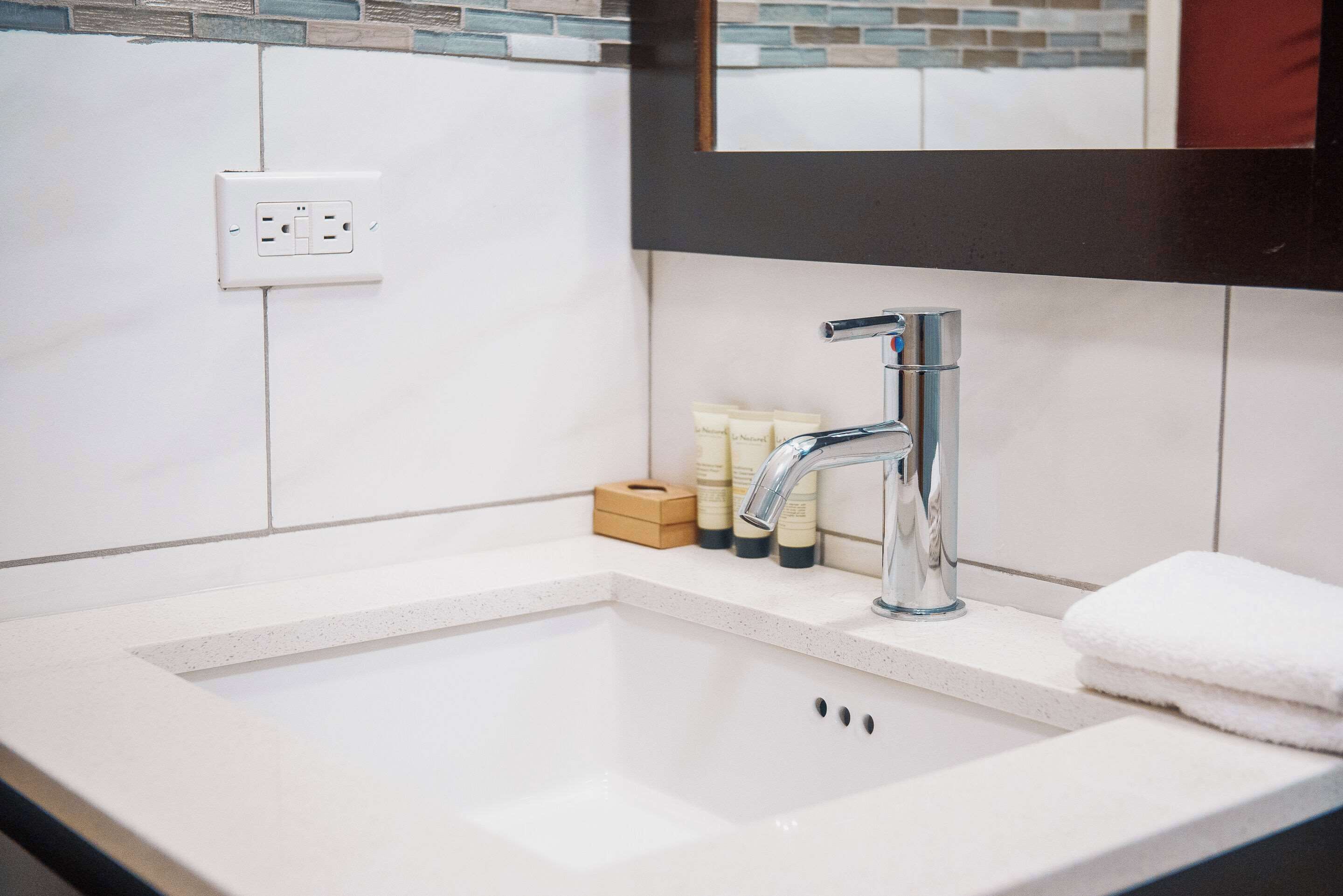 Un bagno con un lavandino bianco, un rubinetto cromato e uno specchio sopra. Ci sono articoli da toilette sul piano di appoggio e una presa elettrica sulla parete.
