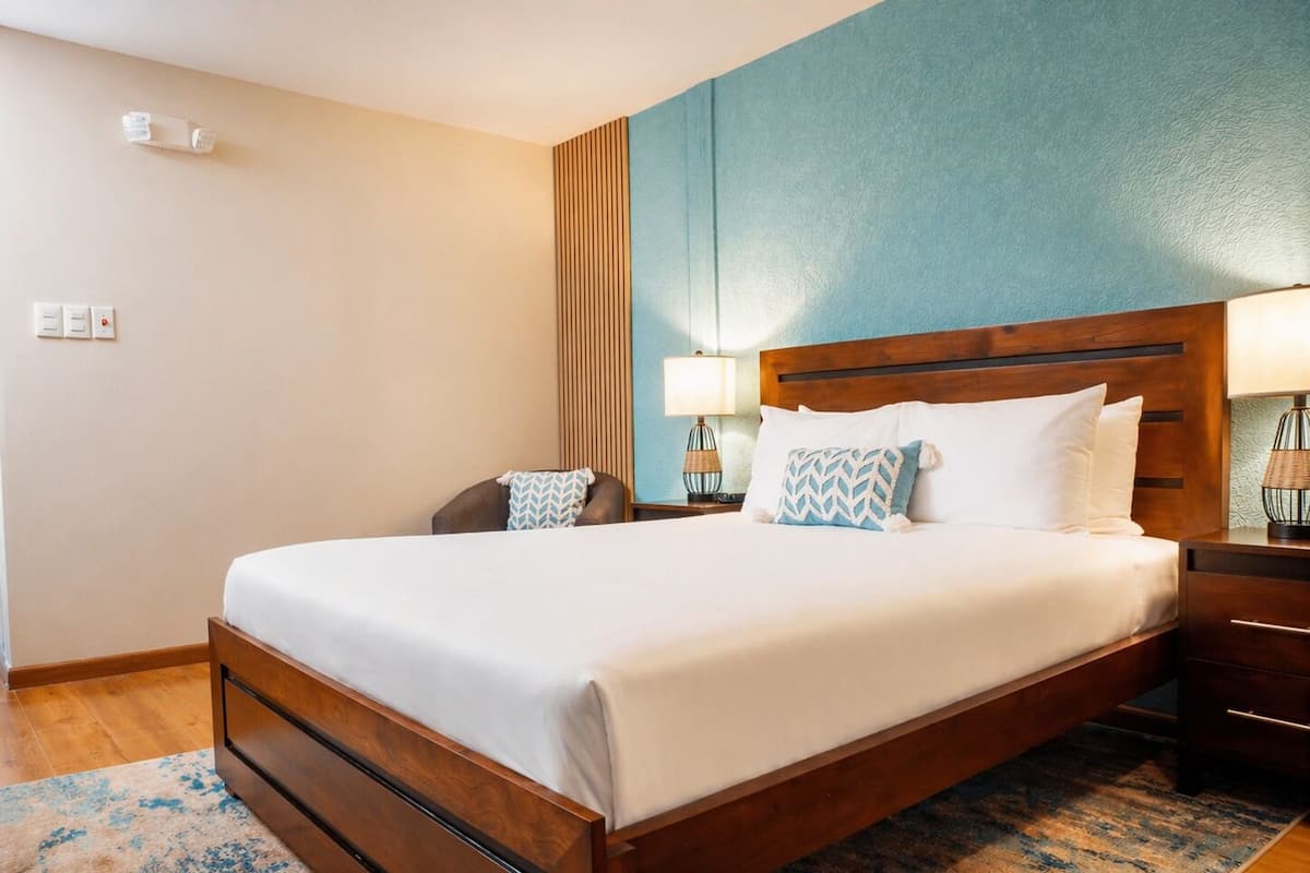 Une chambre d’hôtel avec un grand lit, une tête de lit en bois, deux tables de chevet avec des lampes, une chaise et un tapis à motifs.
