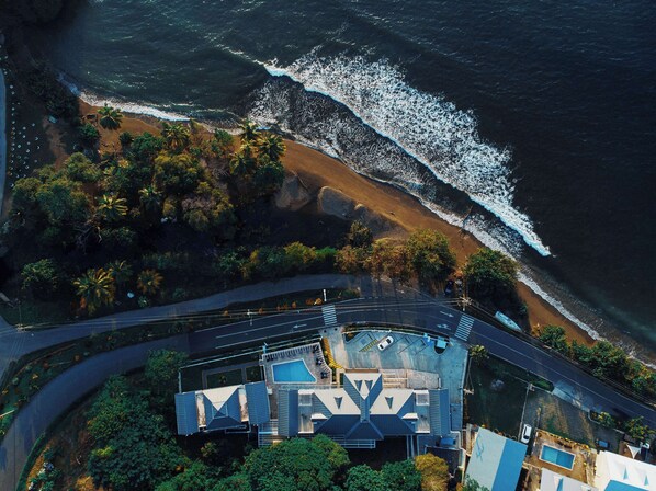 Vista aerea di una proprietà costiera con piscina, area parcheggio e spiaggia, con palme.